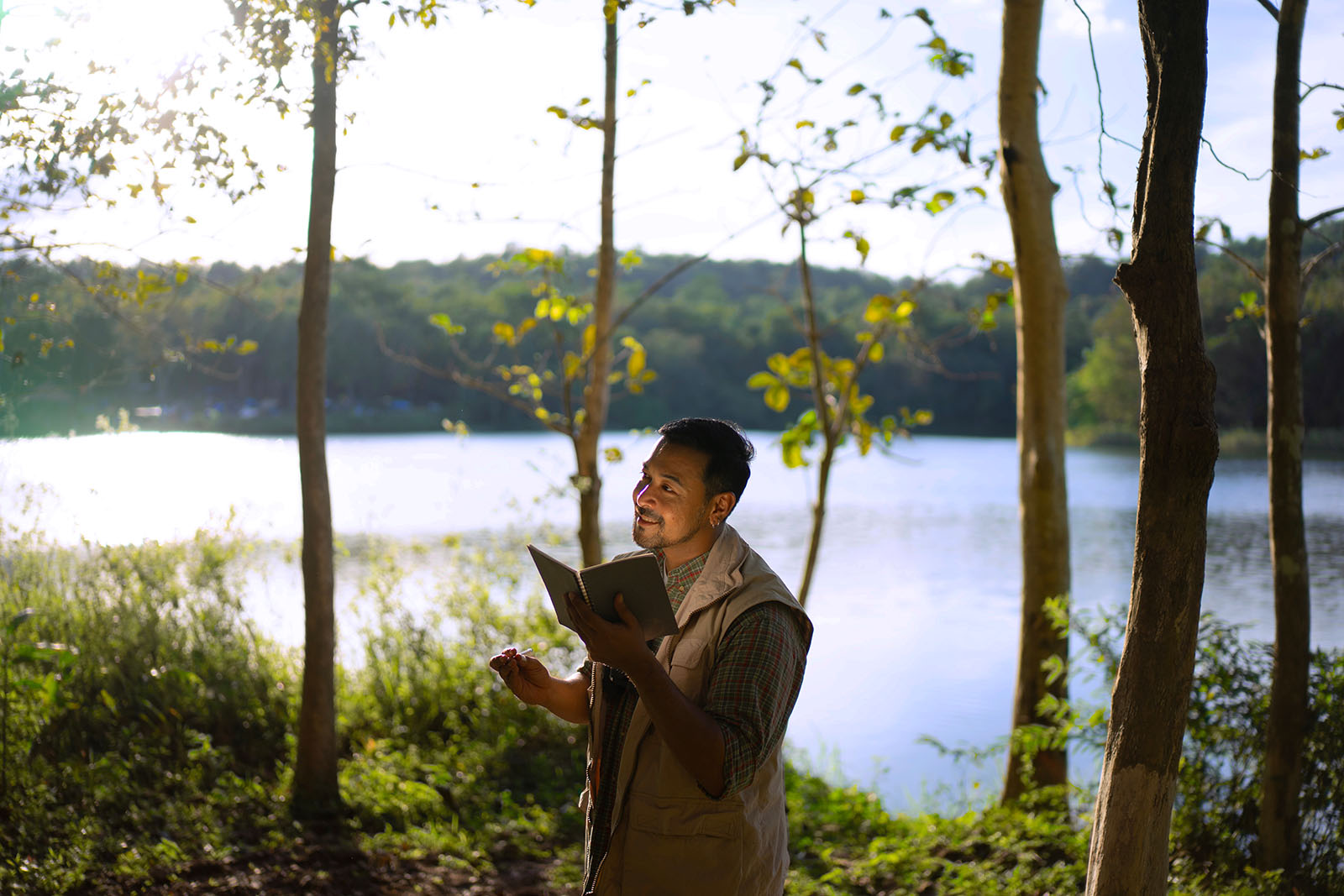 A man in a beige vest and plaid shirt stands near a peaceful lake, surrounded by trees, holding a journal and pen. Sunlight filters through the leaves as he looks up thoughtfully, immersed in nature.